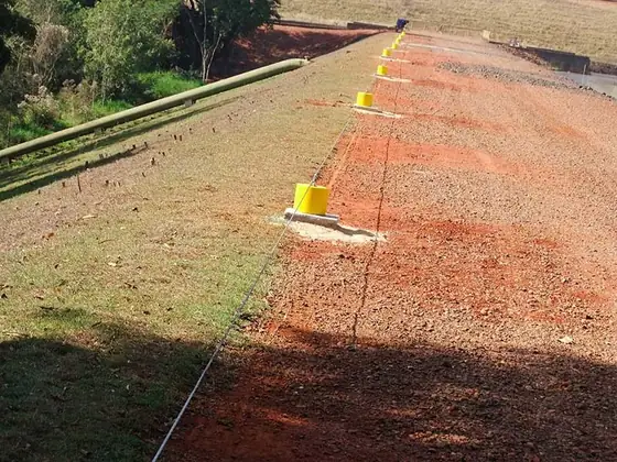 Linha de vida com postes amarelos ao longo de um talude de terra vermelha, em vista longa