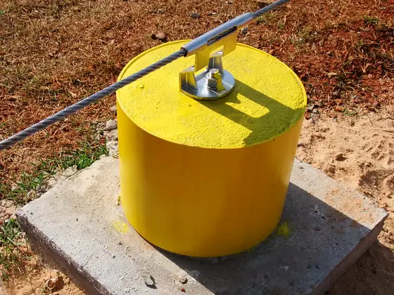 Poste amarelo da linha de vida sobre base de concreto, com ancoragem intermediária no cabo, em solo de terra