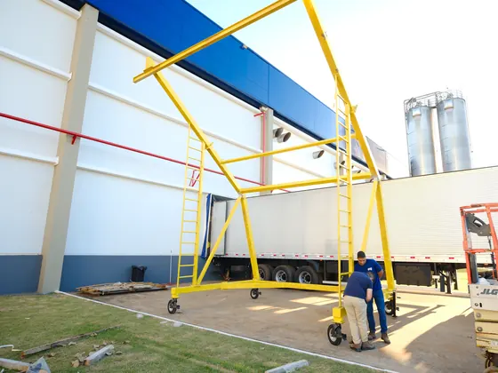 Pórtico móvel C amarelo com escada ao lado de uma carreta, técnicos ao lado