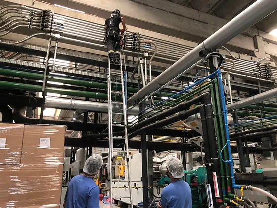Técnico no alto de uma escada móvel entre tubulações de uma fábrica, com dois colegas embaixo